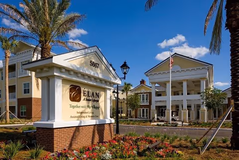 Exterior view of a senior living facility with a large sign reading 'ÉLAN A Senior Lifestyle' and the address 5867. The building features a grand entrance with white columns, an American flag, palm trees, and landscaped flower beds under a blue sky with some clouds.