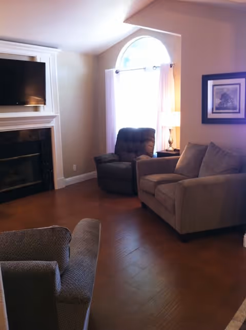Living room with sofa and armchairs facing a fireplace and wall-mounted TV, lit by an arched window.