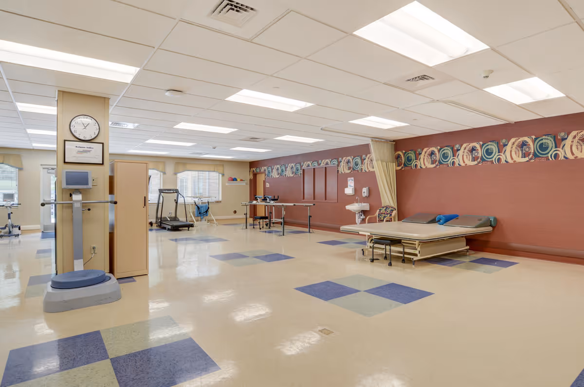 A spacious rehabilitation or therapy room with exercise equipment including a treadmill, parallel bars, and a therapy bed. The room has a tiled floor with blue and green square patterns, a clock on a pillar, and a decorative border along the upper part of one wall. Large windows allow natural light to enter the room.