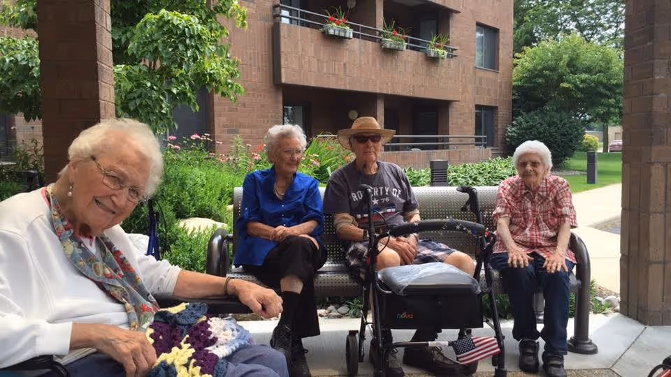Four elderly individuals sitting on benches outside near a brick building with greenery and flowers around. One woman in the foreground is smiling and holding a colorful crocheted blanket. Another woman in a blue shirt, a man wearing a hat and sunglasses, and another woman in a red plaid shirt are seated on a bench behind her. The setting appears to be a peaceful outdoor area of an assisted living community.