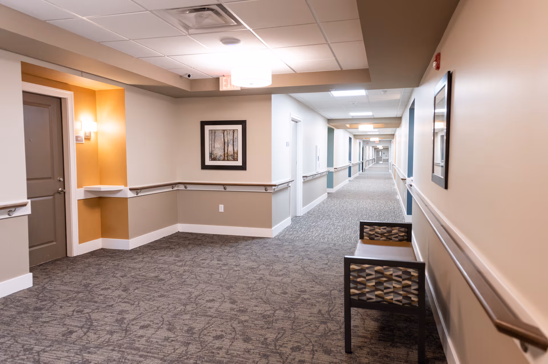 A long, well-lit hallway in a senior living facility with beige walls, carpeted floor, handrails along both sides, framed artwork on the walls, and a patterned chair placed against the right wall near the foreground.