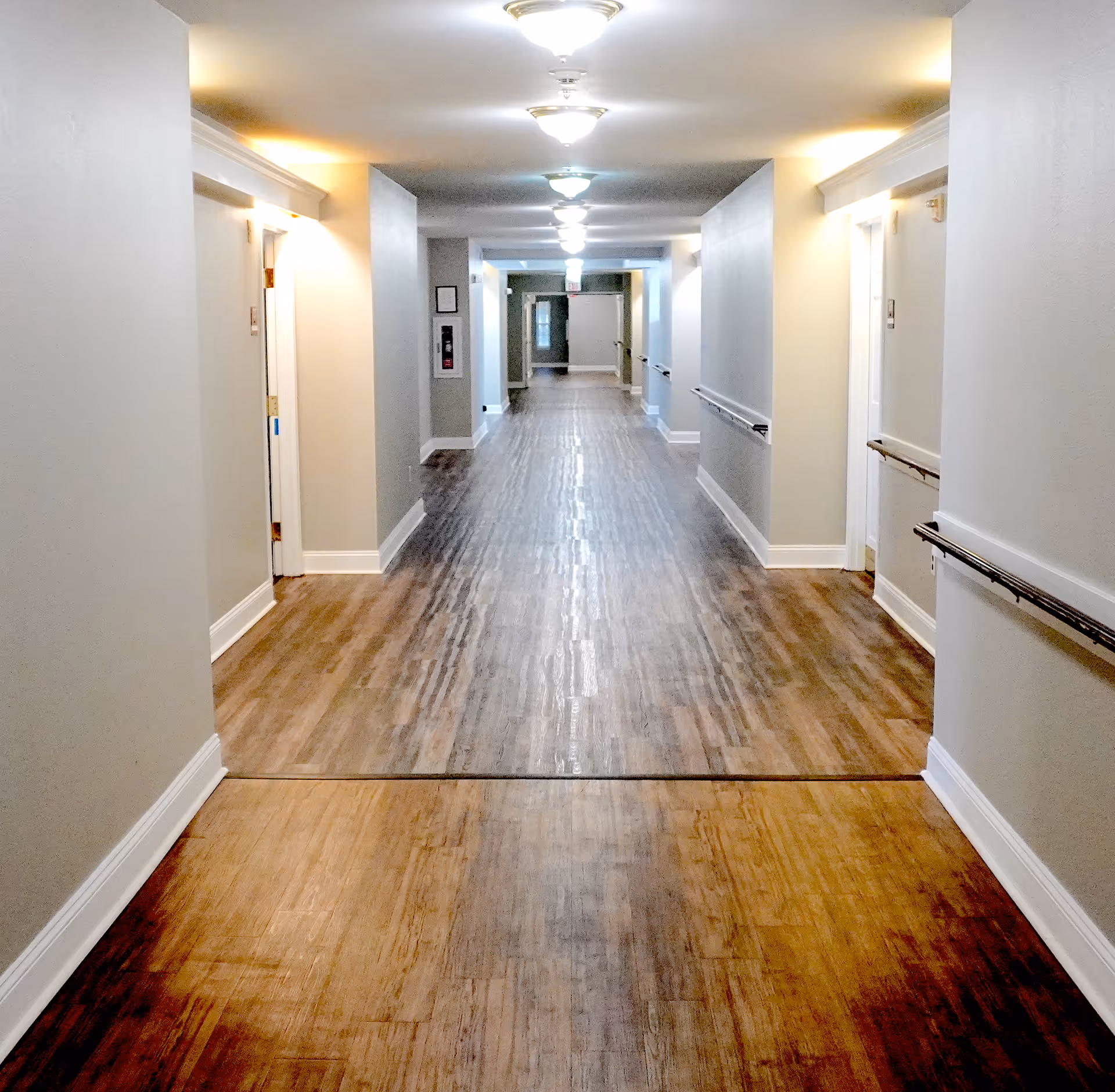 A long, well-lit hallway with wooden flooring and white walls. There are handrails on both sides of the hallway and several doors along the walls. Ceiling lights are evenly spaced along the corridor.