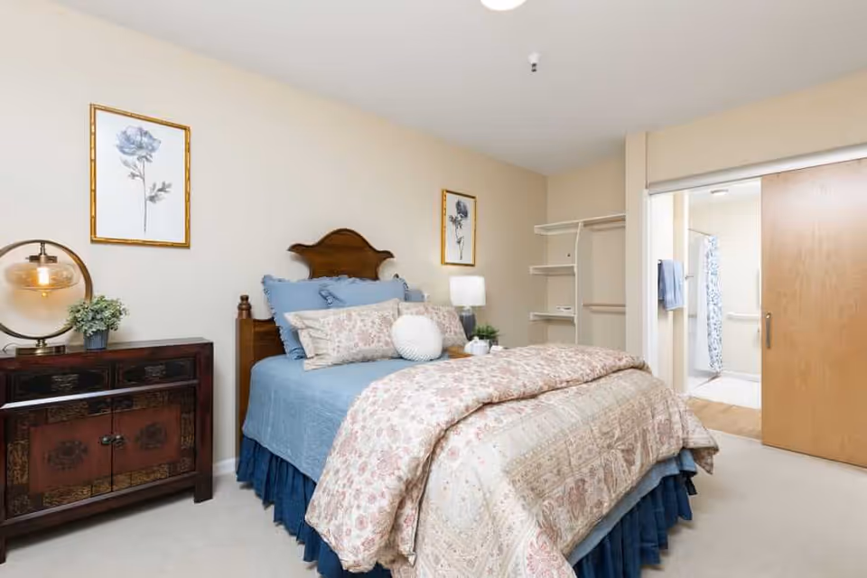 A cozy bedroom in a senior living facility with a wooden headboard bed dressed in blue and floral bedding. There is a wooden nightstand with a round lamp and a small plant on the left side of the bed. Two framed floral artworks hang on the beige walls. An open closet with shelves and hanging space is visible, and a sliding wooden door leads to a bathroom with a shower and towels.