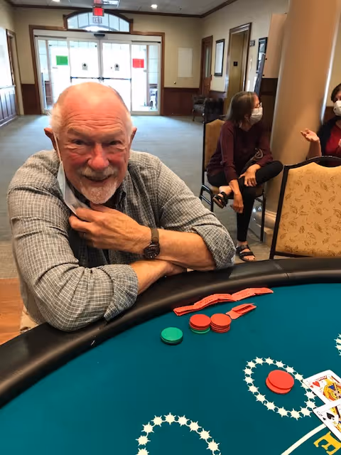 An elderly man with a gray beard and checkered shirt is sitting at a poker table with poker chips and cards in front of him. In the background, two women wearing face masks are seated and engaged in conversation in a well-lit room with large windows and double doors.