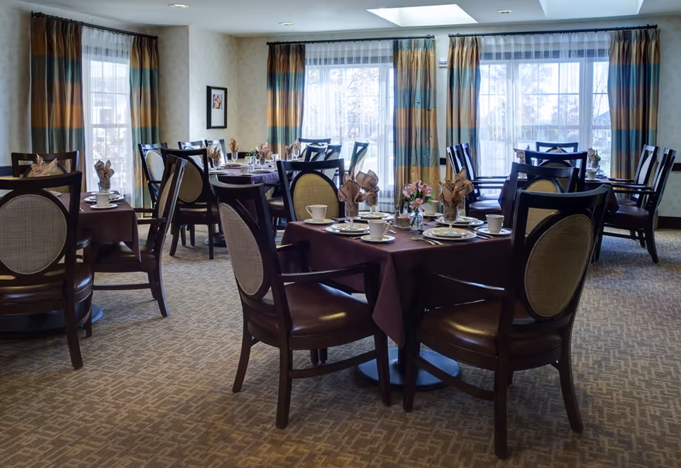 Dining room with multiple round tables set for meals, upholstered chairs, and large windows with patterned curtains.