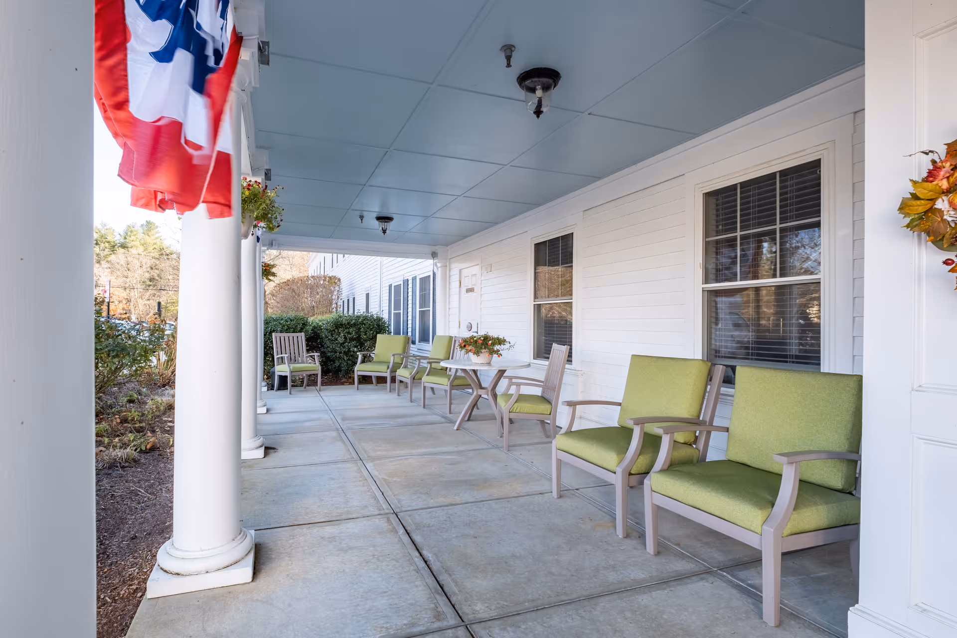 Covered front porch with white columns, green cushioned chairs and small tables outside a senior living facility.