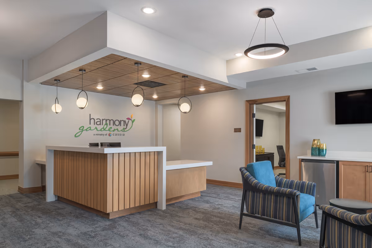 Reception area of Harmony Gardens Senior Living featuring a wooden front desk with white countertop, three hanging globe pendant lights, a seating area with striped and blue cushioned chairs, a wall-mounted TV, and a small cabinet with decorative vases.