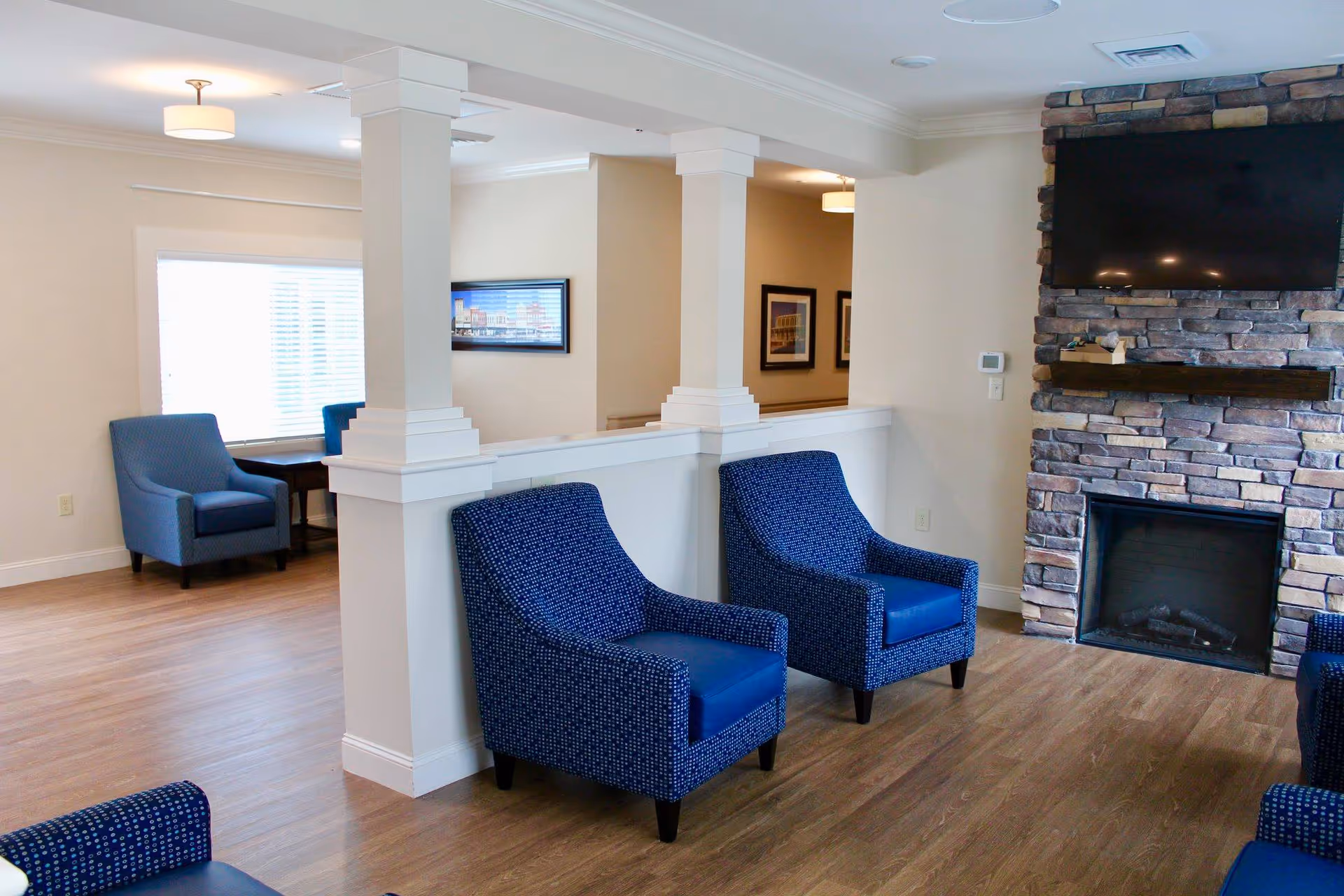 A cozy living room area in Scotland House featuring blue upholstered armchairs arranged near a stone fireplace with a mounted flat-screen TV above it. The room has light-colored walls, wooden flooring, and white pillars dividing the space. In the background, there is a window with blinds and additional seating with a small table.