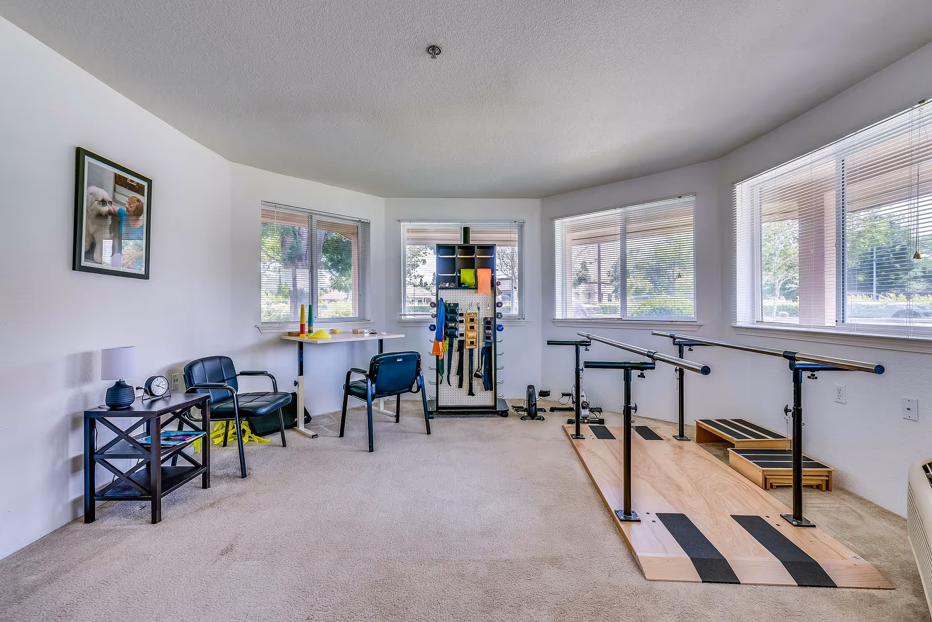 A bright therapy or rehabilitation room with large windows letting in natural light. The room features parallel bars for walking exercises on a wooden platform, two black chairs, a small table with a lamp and clock, and a pegboard with various therapy tools and equipment. A framed picture of a person with a dog hangs on the wall.