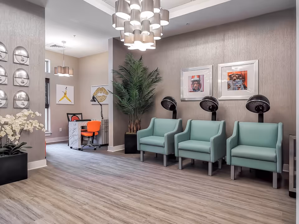 Interior of a senior living facility salon area with three mint green salon chairs under hair dryers, a large potted plant, modern light fixtures, and a small desk with an orange chair in the background. The walls are decorated with framed artwork and metallic face sculptures.