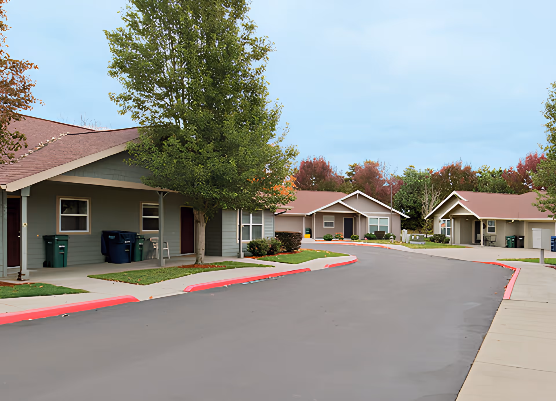 A paved road curves through a senior living community with single-story residential buildings on either side. The buildings have greenish-gray siding and brown roofs. Trees with green and autumn-colored leaves are planted along the sidewalks, and trash bins are visible near the buildings. The sky is clear with a light blue color.