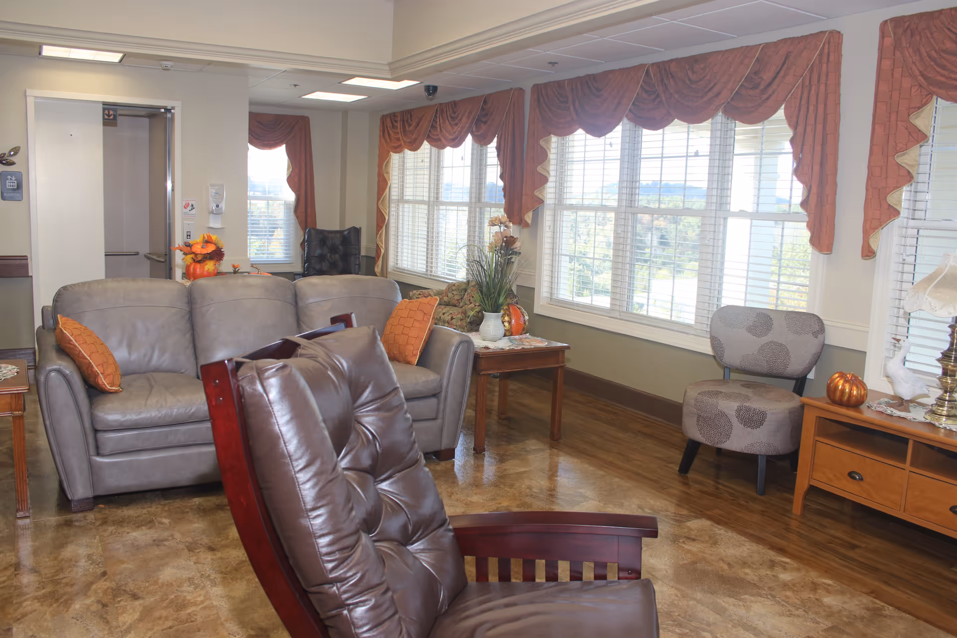 A cozy living room area in a senior living facility with a gray leather sofa, a brown cushioned wooden chair in the foreground, a patterned gray chair, and wooden side tables. Large windows with red valance curtains let in natural light, and decorative items including plants and pumpkins are placed on the tables.