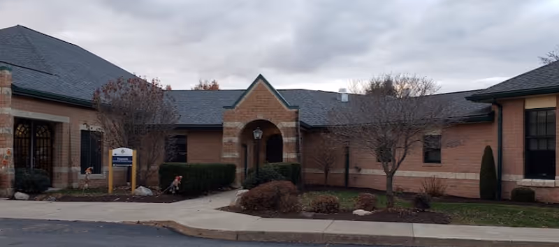 Exterior view of a single-story brick building with a peaked roof and an arched entrance. There are leafless trees and shrubs in front of the building, and a sidewalk leading to the entrance. The sky is cloudy.