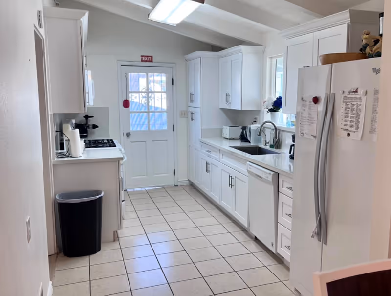 Bright narrow kitchen with white cabinets, tiled floor, sink and dishwasher on the right, stove and trash can on the left, and a door at the far end.