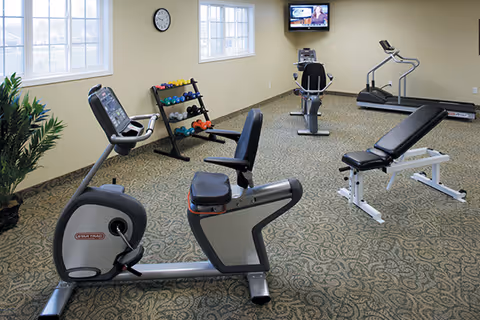 A fitness room with exercise equipment including a recumbent stationary bike, a treadmill, a weight bench, a stationary bike, and a rack of colorful dumbbells. The room has beige walls, two windows, a clock, a wall-mounted television, and patterned carpet flooring.