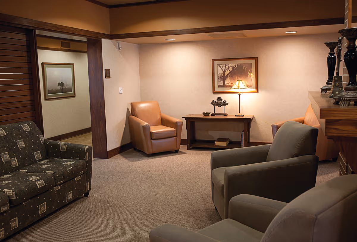 A cozy senior living facility common area with several armchairs and a patterned sofa arranged around a wooden table with a lamp and decorative items. The walls are beige with framed artwork, and the room has warm lighting and carpeted flooring.