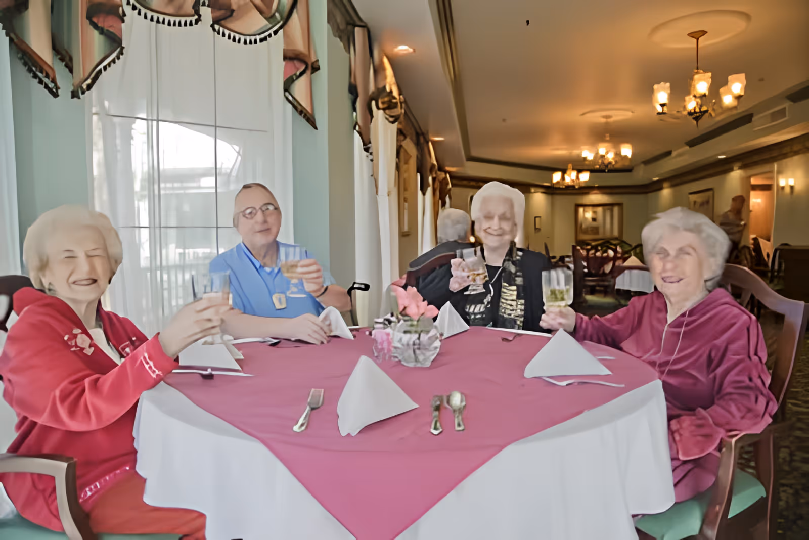 Four elderly residents sit around a round table in a dining room, smiling and raising glasses in a toast.