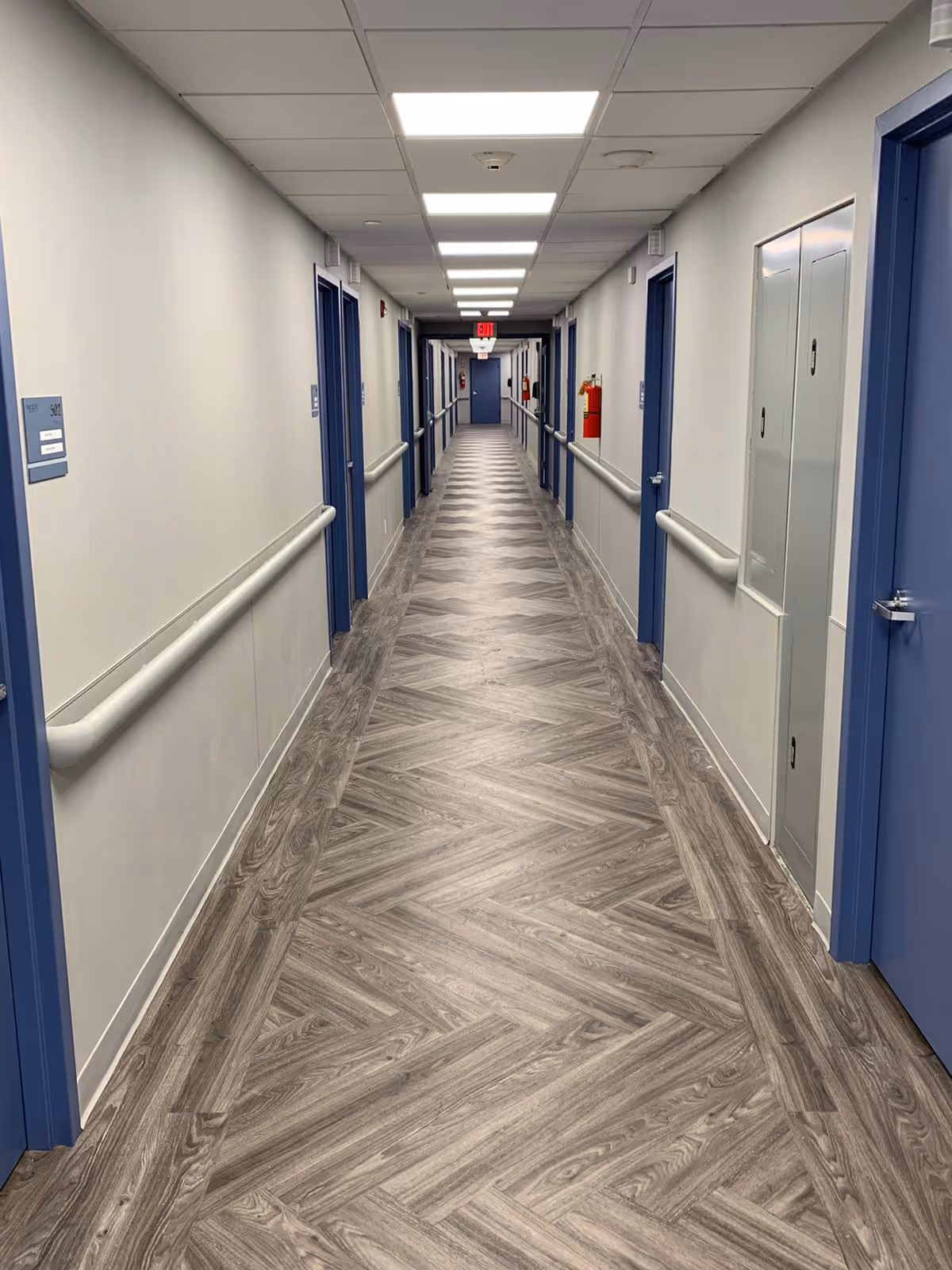 Long indoor hallway with wood-patterned flooring and white walls, lined with blue doors on both sides. Handrails run along the walls, and ceiling lights illuminate the corridor. A red fire extinguisher is mounted on the right wall, and an exit sign is visible at the far end.