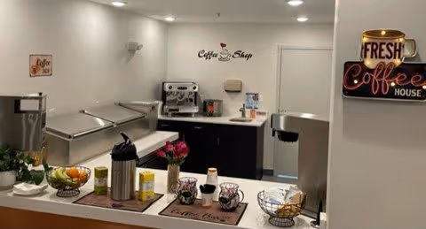 Interior view of a coffee shop area in a senior living facility with a coffee machine, coffee supplies, fruit bowl, and decorative signs on the walls reading 'Coffee Shop' and 'Fresh Coffee House'.