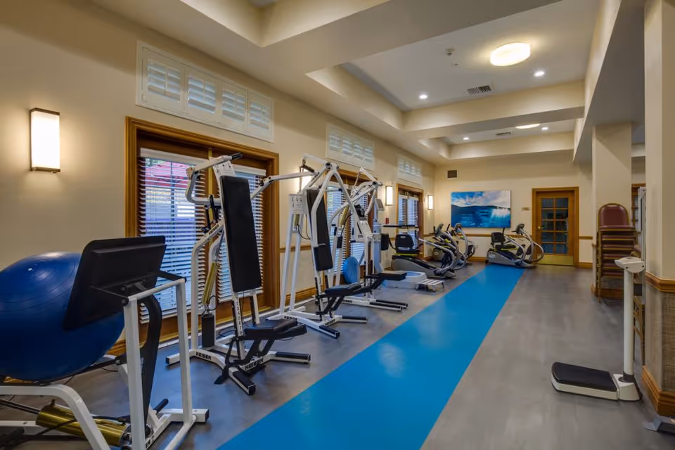 A fitness room in a senior living facility with various exercise machines including weight machines, stationary bikes, and a large blue exercise ball. The room has wooden framed windows with blinds, beige walls, and a blue exercise mat running along the floor. There is a painting of an ocean wave on the far wall and a stack of chairs near a door.