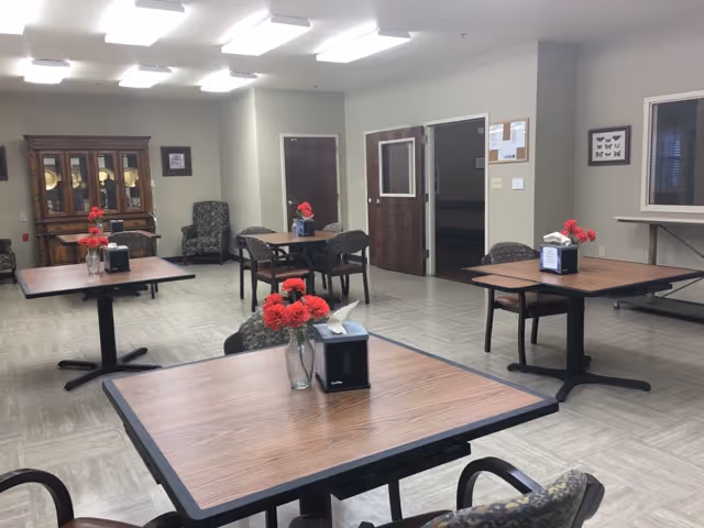 A dining room in a senior living facility with several square wooden tables, each decorated with a vase of red flowers and a napkin dispenser. The room has beige walls, a wooden cabinet with glass doors, patterned chairs, and fluorescent ceiling lights. There are doors and windows visible on the far wall.