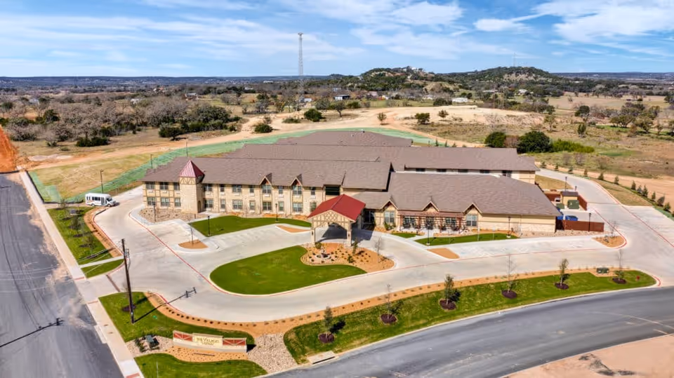 Aerial view of The Villages of Windcrest senior living facility showing a large, two-story building with a brown roof and stone exterior. The building is surrounded by paved driveways, landscaped green lawns, and small trees. The surrounding area includes open land and distant hills under a partly cloudy sky.