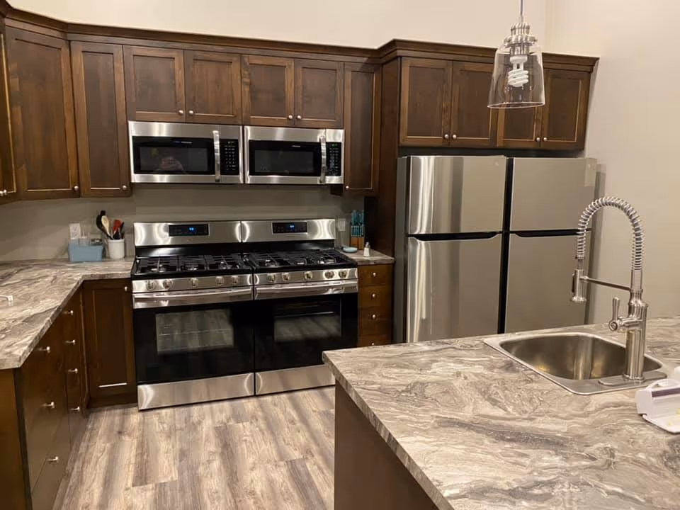 Modern kitchen with dark wooden cabinets, two stainless steel microwaves above a double oven stove, a double-door stainless steel refrigerator, and a marble countertop island with a sink and a flexible spring faucet. The floor has a light wood finish.