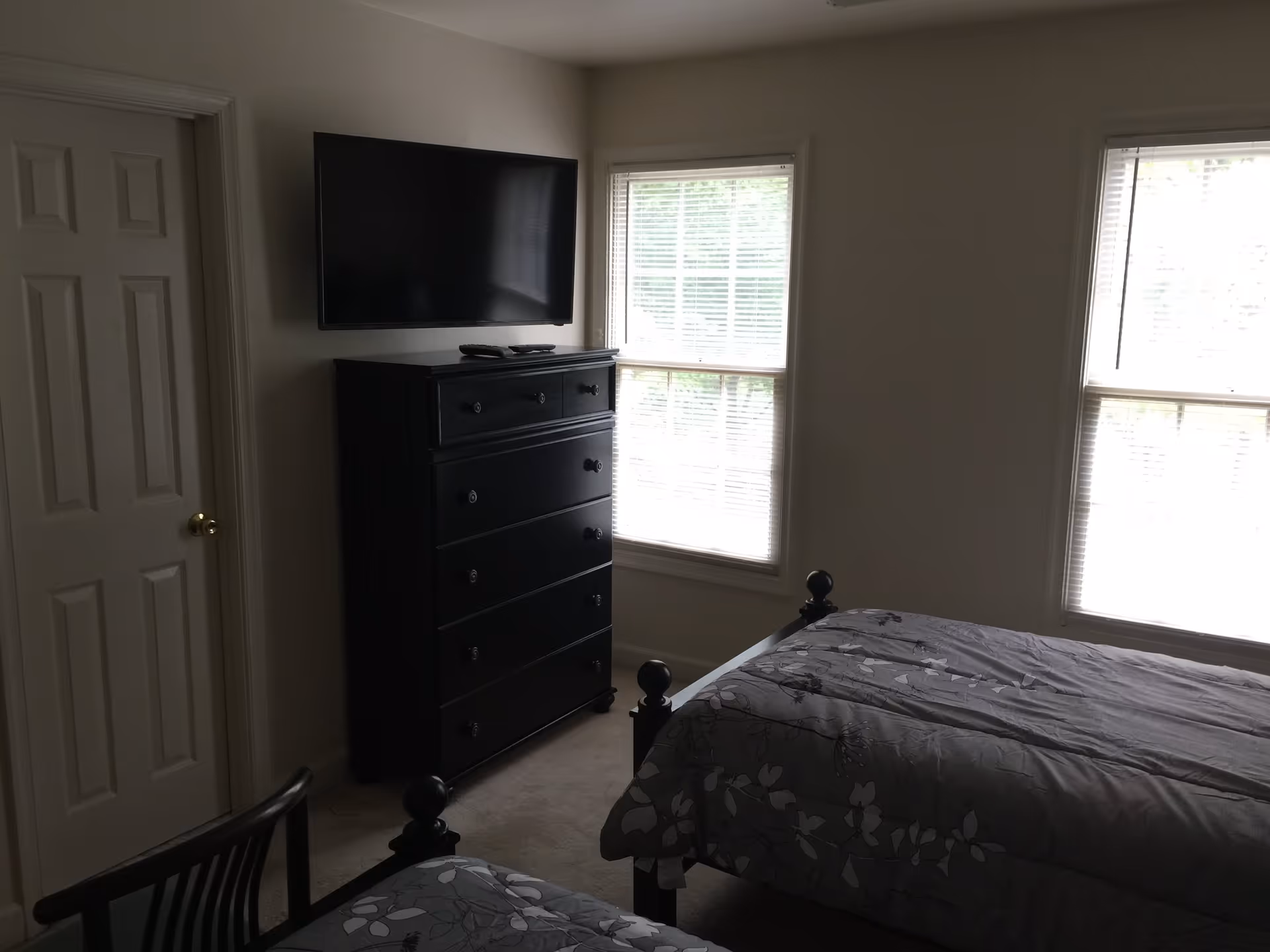A bedroom with two beds covered in gray floral-patterned bedding, a black dresser with a flat-screen TV mounted above it, two large windows with blinds, a closed door, and a wooden chair partially visible in the foreground.