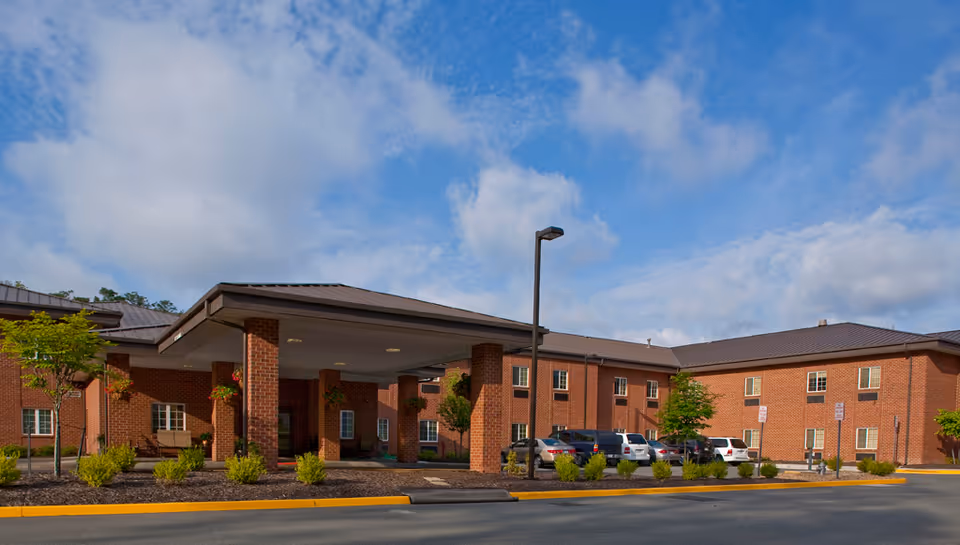 Exterior view of a two-story brick senior living facility building with a covered entrance, several parked cars, small trees, and shrubs under a partly cloudy blue sky.