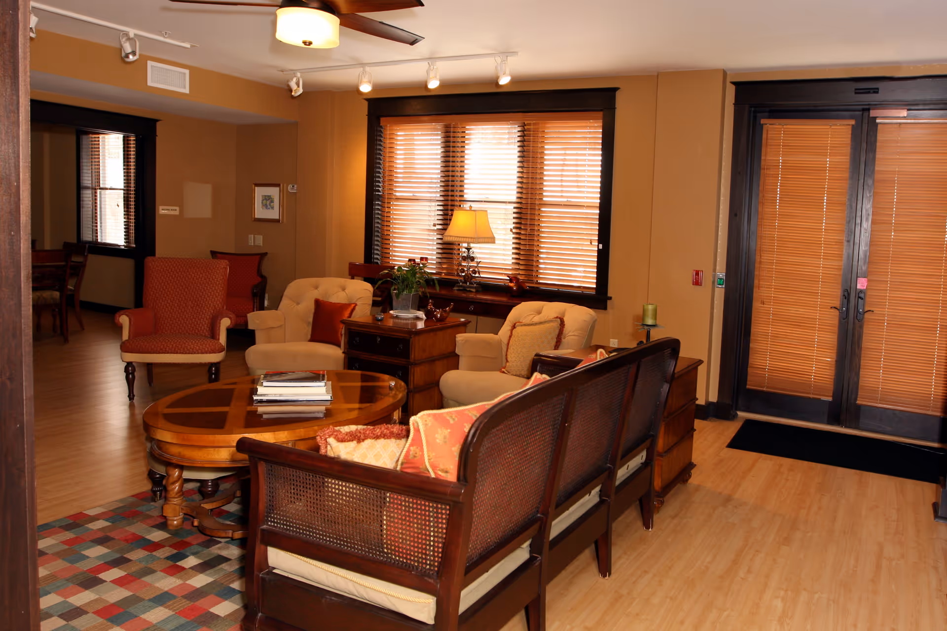 A cozy common living room with upholstered chairs, a wooden coffee table, sofa seating, and windows with wooden blinds near double doors.