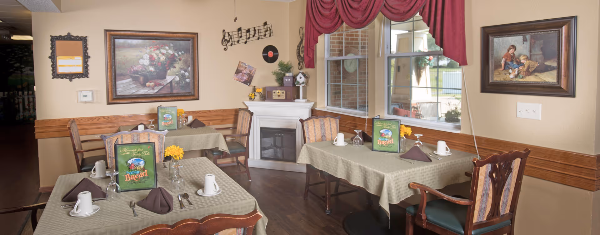 Cozy dining room with several tables set with cups and folded napkins, wooden chairs, wall paintings, and a small decorative fireplace beneath a window.