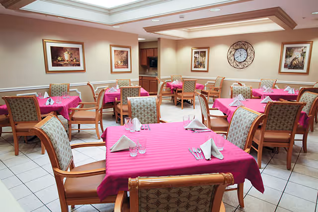 A dining room with multiple tables covered in bright pink tablecloths, each set with folded white napkins, glasses, and silverware. The room has beige walls adorned with framed artwork and a large decorative clock. Wooden chairs with patterned upholstery surround the tables, and the floor is tiled in a light color.