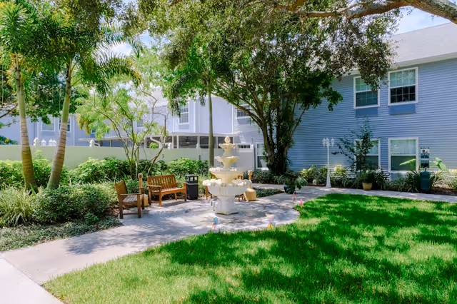 Outdoor courtyard area with a multi-tiered white fountain in the center, surrounded by green grass, trees, and shrubs. There are wooden benches and a trash can along a paved walkway. A light blue building with white trim and windows is visible in the background.