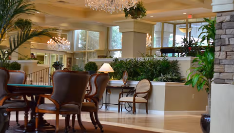 Spacious, well-lit communal dining and lounge area with tables, upholstered chairs, plants, and chandeliers.