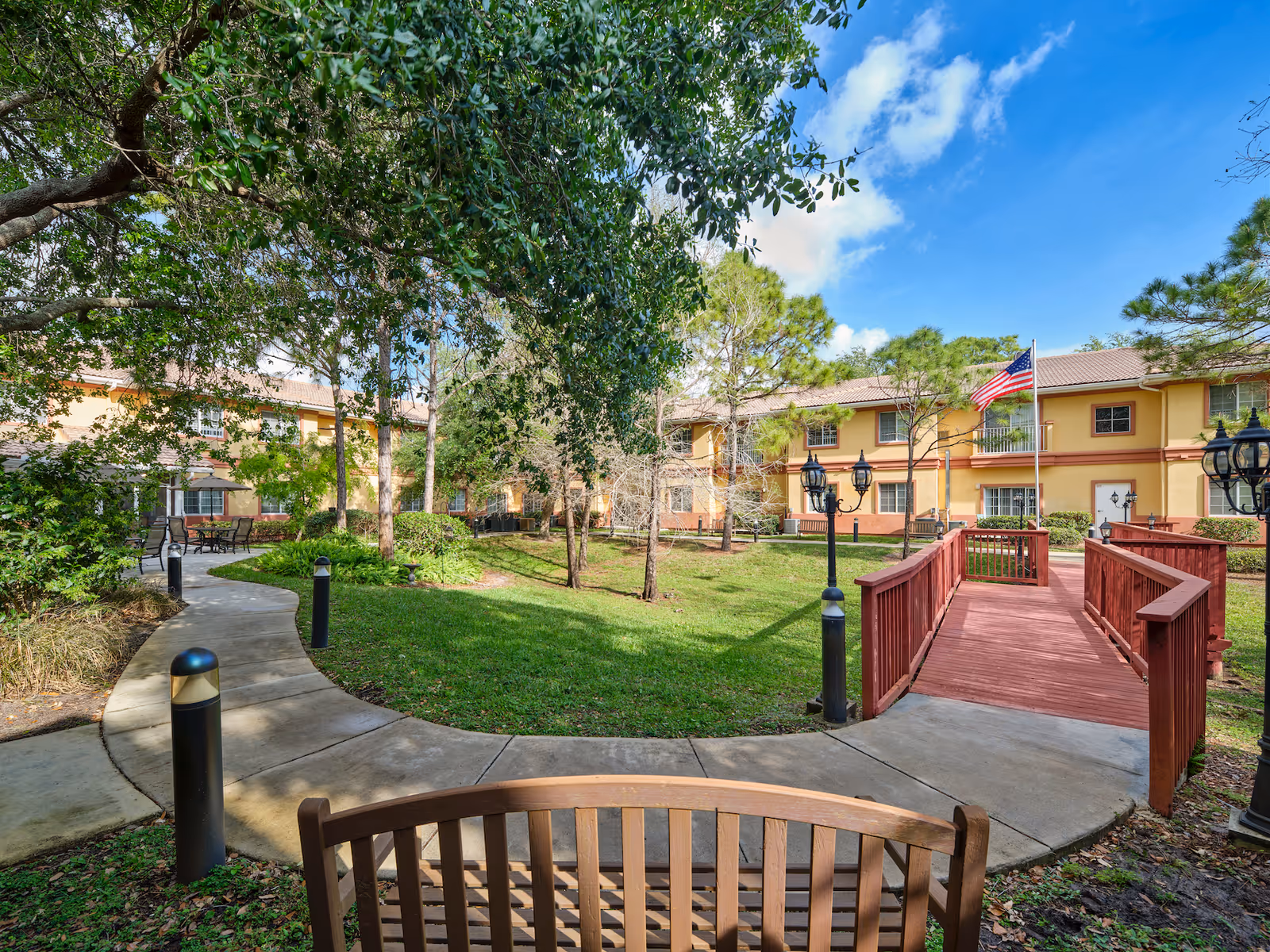 Curved garden courtyard with a wooden bench and red footbridge leading to a two-story yellow building surrounded by trees and a blue sky.