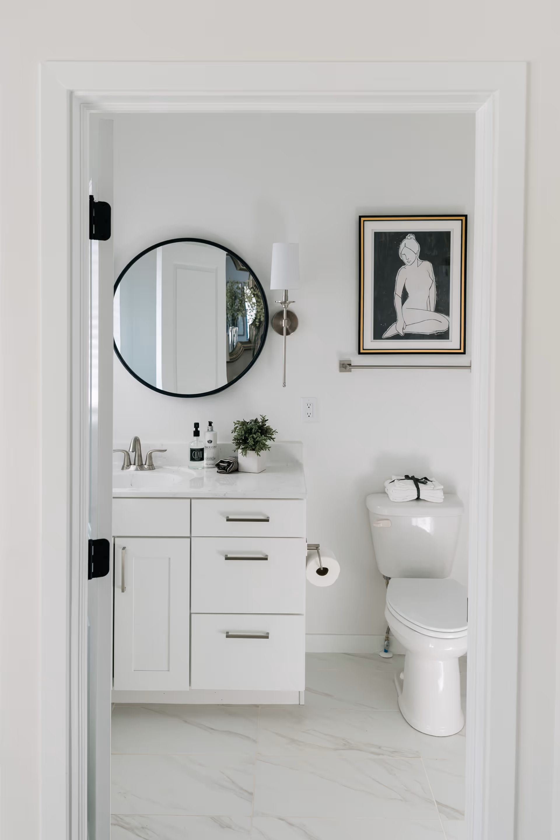 White bathroom with a vanity and round mirror, wall sconce, framed artwork, and a toilet.