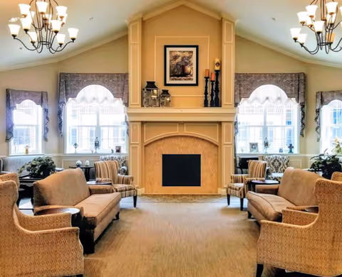 A spacious and well-lit living room area with a central fireplace. The room features two chandeliers, large windows with decorative curtains, and multiple upholstered chairs and sofas arranged symmetrically around the fireplace. The fireplace mantle is decorated with candles and framed artwork above it.