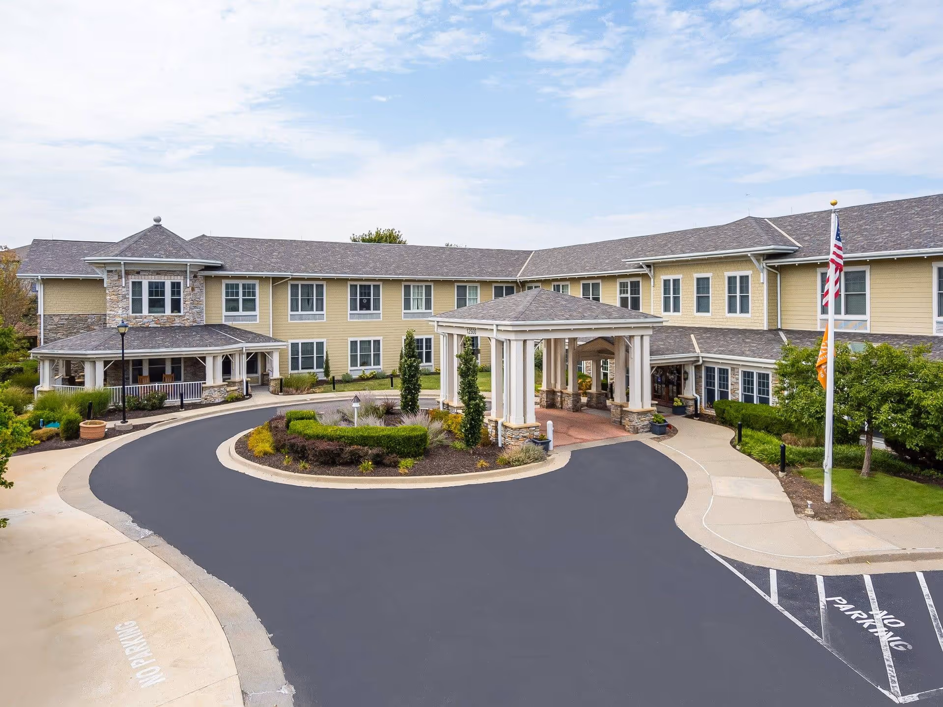 Exterior view of Sunrise of Overland Park senior living facility showing a two-story building with beige siding and stone accents, a covered entrance with white columns, a circular driveway, landscaped greenery, and an American flag on a flagpole.