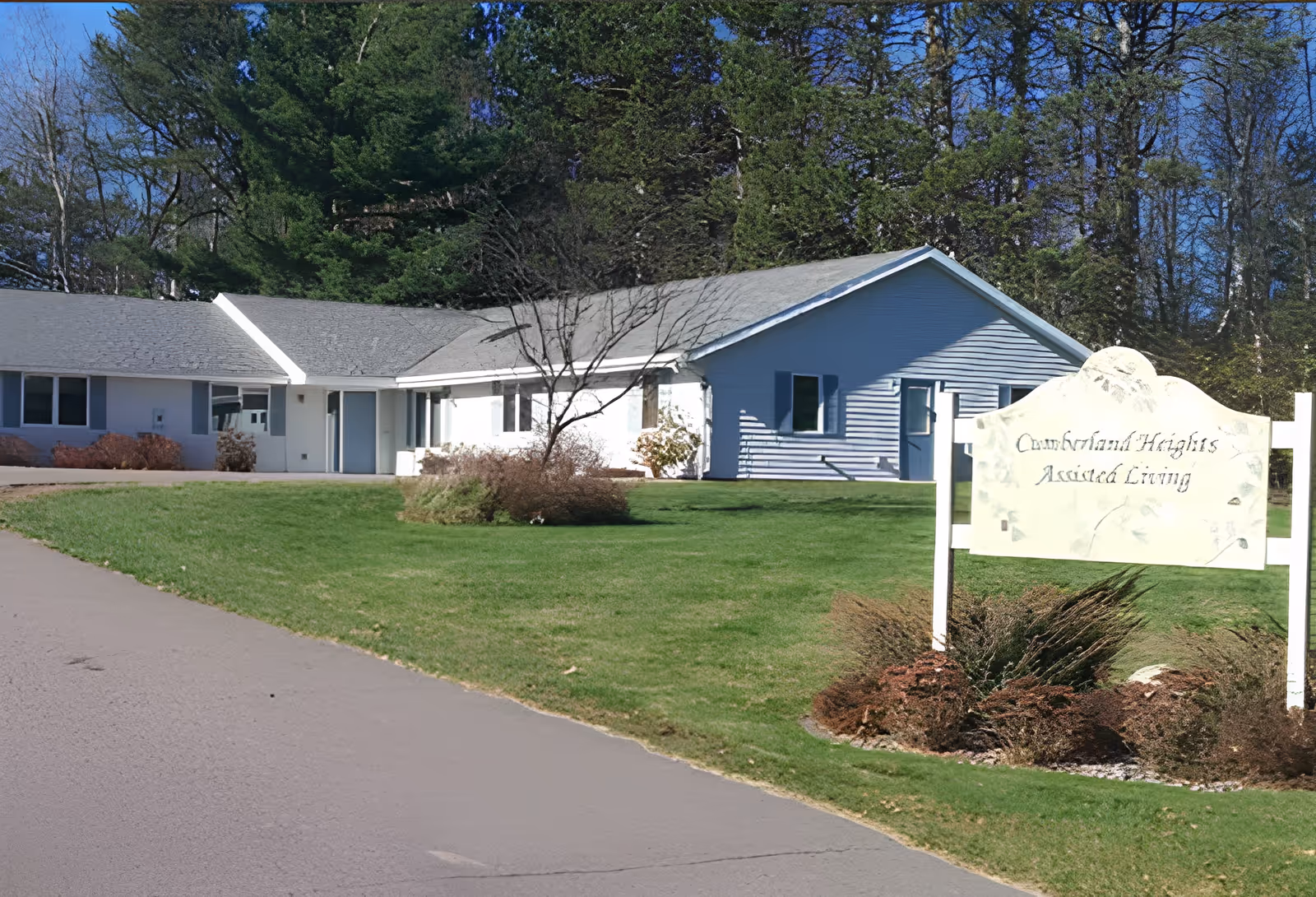 Exterior view of a single-story building with a gray roof and white siding, surrounded by green grass and trees. In the foreground, there is a sign that reads 'Cumberland Heights Assisted Living'.