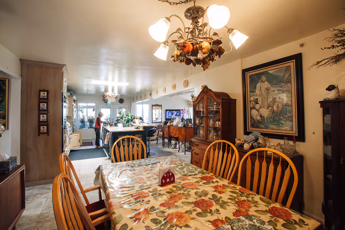 Dining room area with a floral tablecloth-covered table and wooden chairs, a chandelier overhead, a china cabinet, and a kitchen visible in the background.
