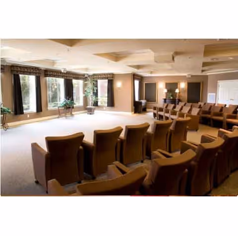 A spacious room with multiple rows of brown cushioned chairs arranged facing a blank wall, large windows with dark curtains letting in natural light, and potted plants near the windows. The room has a beige carpet and neutral-colored walls with ceiling light fixtures.