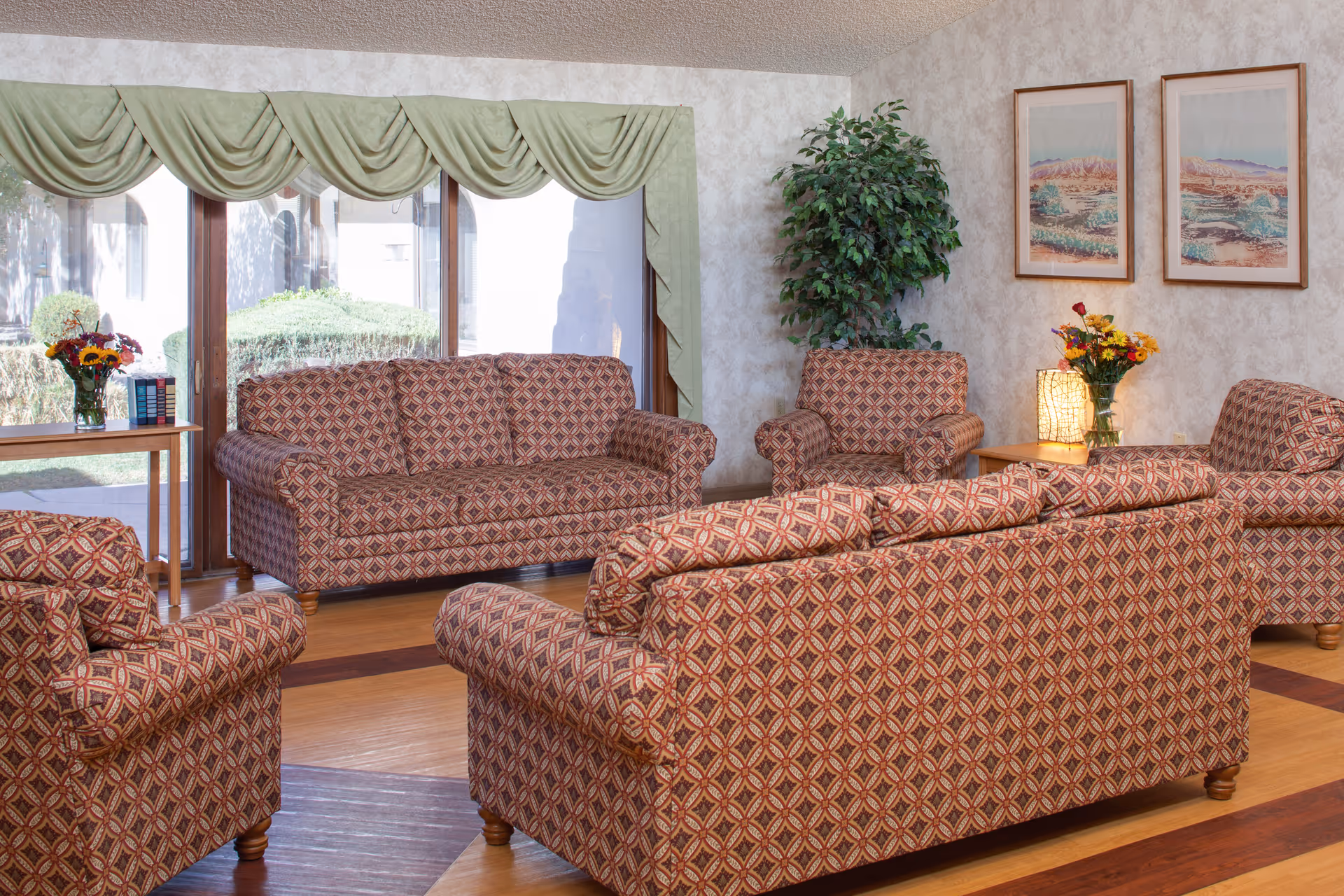 A living room area with patterned upholstered sofas and armchairs arranged around a wooden floor. There is a large window with green drapes, a side table with a vase of flowers and books, a potted plant in the corner, and two framed landscape paintings on the wall.