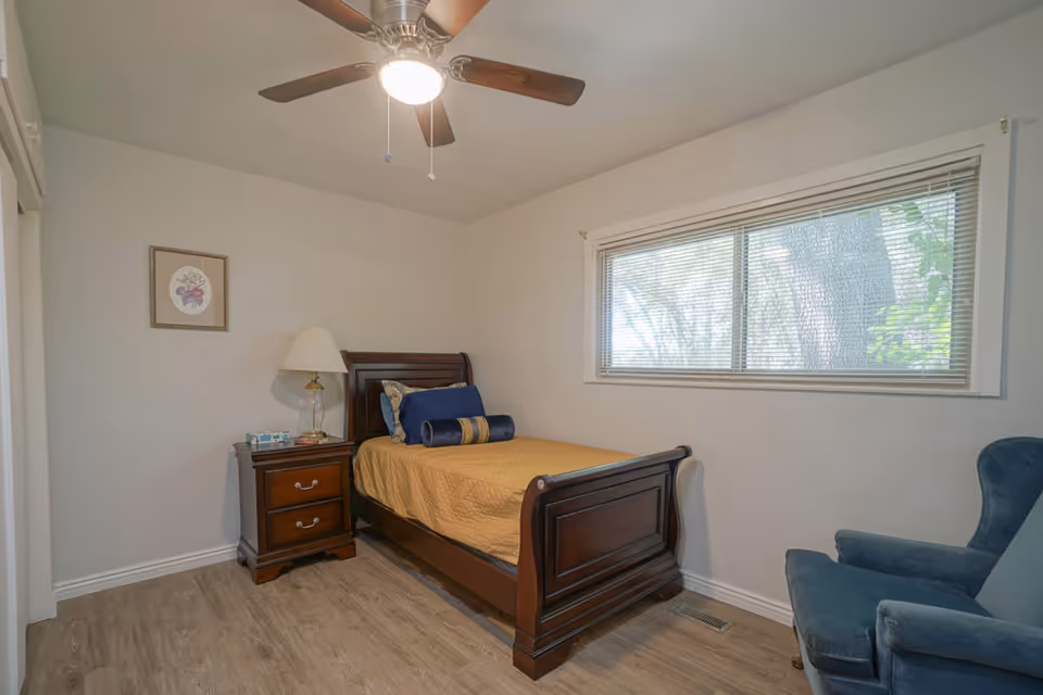 A small bedroom with a single wooden bed covered with a yellow bedspread and blue pillows. Next to the bed is a wooden nightstand with a glass lamp and some items on it. A framed picture hangs on the wall above the nightstand. There is a large window with blinds on the right wall and a blue upholstered armchair in the corner. The ceiling has a fan with a light turned on.