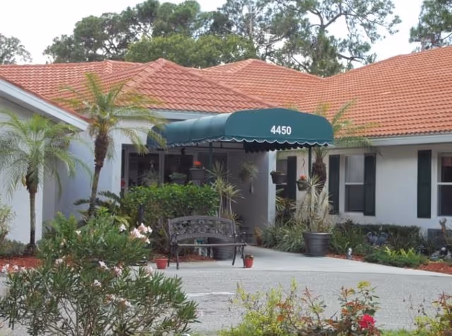Exterior view of a single-story building with a red tiled roof and white walls. The entrance has a green awning with the number 4450 on it. There are palm trees, various plants, and flowers around the entrance, along with a black metal bench on the walkway.