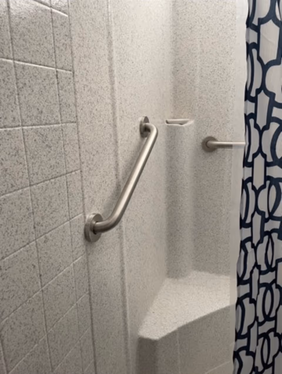 Shower area with textured light-colored walls, a built-in corner seat, a metal grab bar on the wall, and a shower curtain with a blue and white geometric pattern.