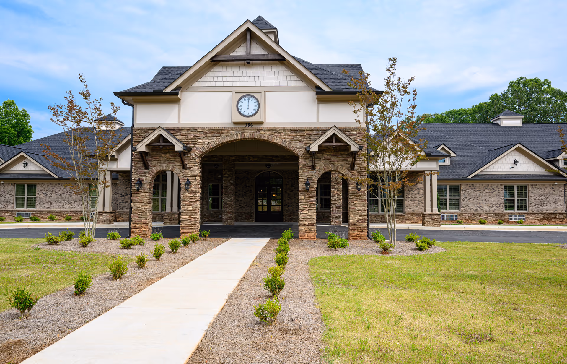 Front view of a senior living facility with a stone-covered entrance portico, clock above, and a walkway leading to the doors.