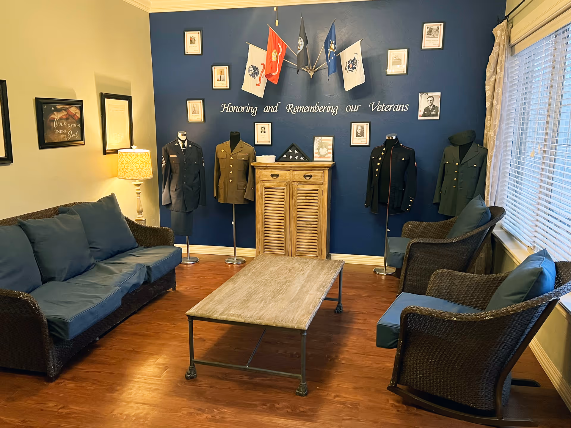 A cozy living room area with a blue accent wall displaying military uniforms on mannequins, framed photos, and flags. The wall has the words 'Honoring and Remembering our Veterans' written on it. The room has a wicker sofa and two wicker armchairs with blue cushions, a wooden coffee table, a small wooden cabinet, a lamp, and window blinds letting in natural light.