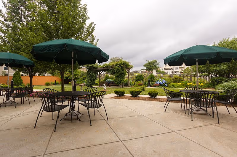 Outdoor patio area with several round metal tables and chairs, each table shaded by a large green umbrella. The patio is surrounded by well-maintained landscaping including bushes, trees, and flowering plants. In the background, there is a garden archway and a fence with buildings visible beyond it under a cloudy sky.