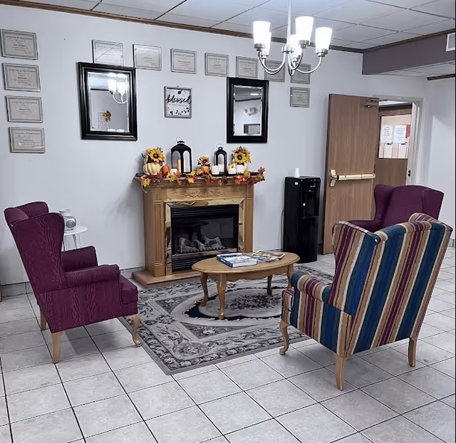 A cozy sitting area in a senior living facility with a wooden fireplace decorated with autumn-themed items including sunflowers, pumpkins, and candles. Two purple armchairs and one striped armchair surround a small wooden coffee table on a patterned rug. The wall behind the fireplace features framed certificates, two mirrors, and a decorative sign that says 'blessed'. A chandelier with five lights hangs from the ceiling, and a water cooler is visible near an open door.