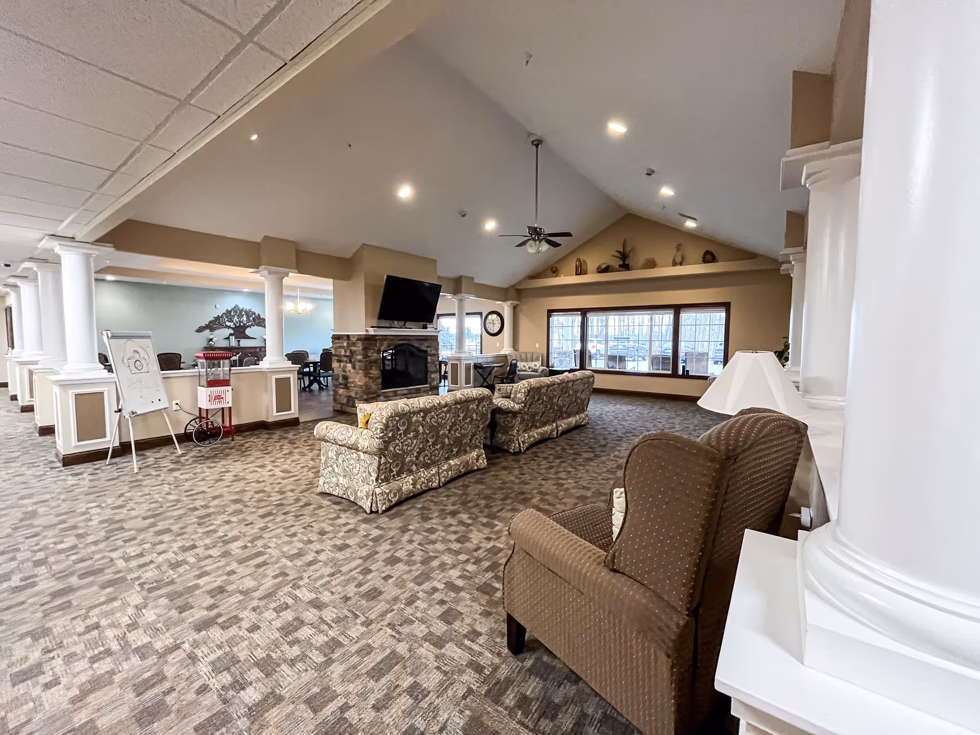 Spacious senior living facility common area with patterned carpet, multiple upholstered chairs and sofas arranged around a stone fireplace with a mounted flat-screen TV. Large windows let in natural light, and white columns separate the seating area from a dining space with tables and chairs. The ceiling is vaulted with recessed lighting and a ceiling fan.